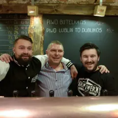 Trzech uśmiechniętych mężczyzn za **tap barem** w pubie. Na tablicy za nimi widnieje menu piwne.