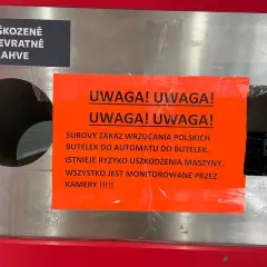 Na automacie do butelek wisi pomarańczowa tabliczka z napisem "UWAGA! UWAGA! SUROWY ZAKAZ WRZUCANIA POLSKICH BUTELEK DO AUTOMATU DO BUTELEK. ISTNIEJE RYZYKO USZKODZENIA MASZYNY. WSZYSTKO JEST MONITOROWANE PRZEZ KAMERY !!!!!". Obok znajduje się czarna t...