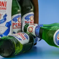 Butelki piwa Peroni Nastro Azzurro leżą na niebieskim tle. Jedna z butelek jest otwarta, a pianka unosi się z szyjki.