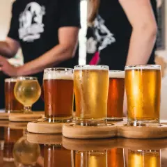 Degustacja piwa typu ale. Kilka szklanek z bursztynowym i złotym piwem na drewnianych podstawkach, odbijających się w lustrzanym blacie stołu.