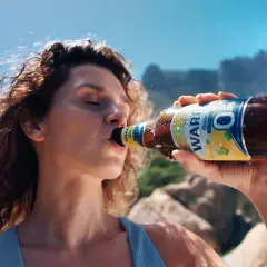 Kobieta pije piwo Warka Radler 0,0% na plaży. Idealne na upalne dni, gdy chcesz orzeźwienia bez procentów.