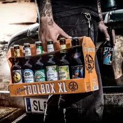 Mężczyzna z tatuażami niesie skrzynkę "TOOLBOX XL" z różnymi butelkami piwa. Idealne połączenie: jakie butelki do jakich skrzynek.