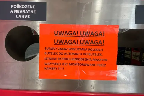 Na automacie do butelek wisi pomarańczowa tabliczka z napisem "UWAGA! UWAGA! SUROWY ZAKAZ WRZUCANIA POLSKICH BUTELEK DO AUTOMATU DO BUTELEK. ISTNIEJE RYZYKO USZKODZENIA MASZYNY. WSZYSTKO JEST MONITOROWANE PRZEZ KAMERY !!!!!". Obok znajduje się czarna t...