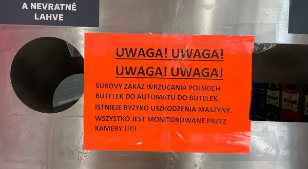 Na automacie do butelek wisi pomarańczowa tabliczka z napisem "UWAGA! UWAGA! SUROWY ZAKAZ WRZUCANIA POLSKICH BUTELEK DO AUTOMATU DO BUTELEK. ISTNIEJE RYZYKO USZKODZENIA MASZYNY. WSZYSTKO JEST MONITOROWANE PRZEZ KAMERY !!!!!". Obok znajduje się czarna t...
