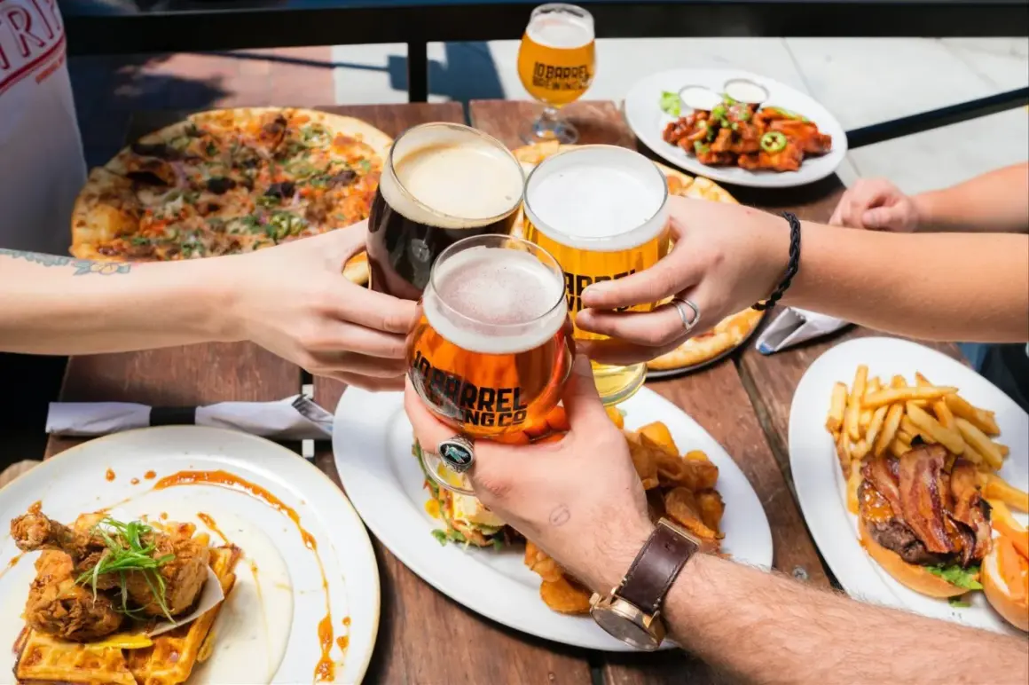 Ludzie wznoszą toasty piwem, celebrując spożycie piwa na świecie przy stole pełnym pizzy, burgerów i kurczaka z goframi.