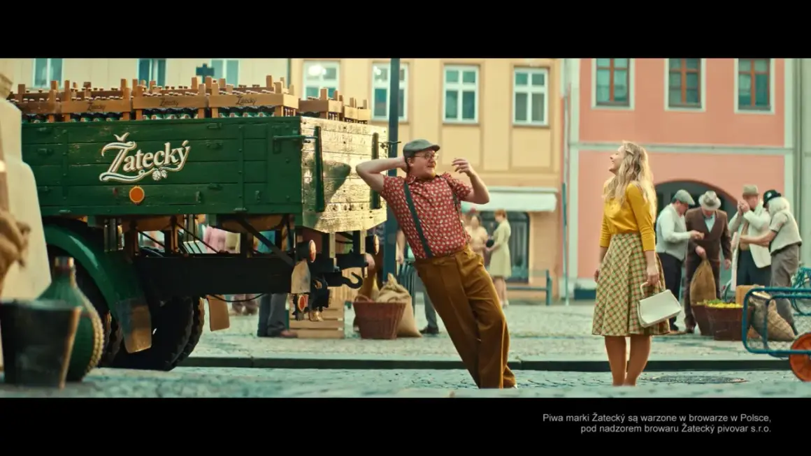 Mężczyzna w czapce i szelkach opiera się o ciężarówkę z piwem Žatecký. Ile procent ma to piwo? Kobieta w żółtej bluzce patrzy w górę.