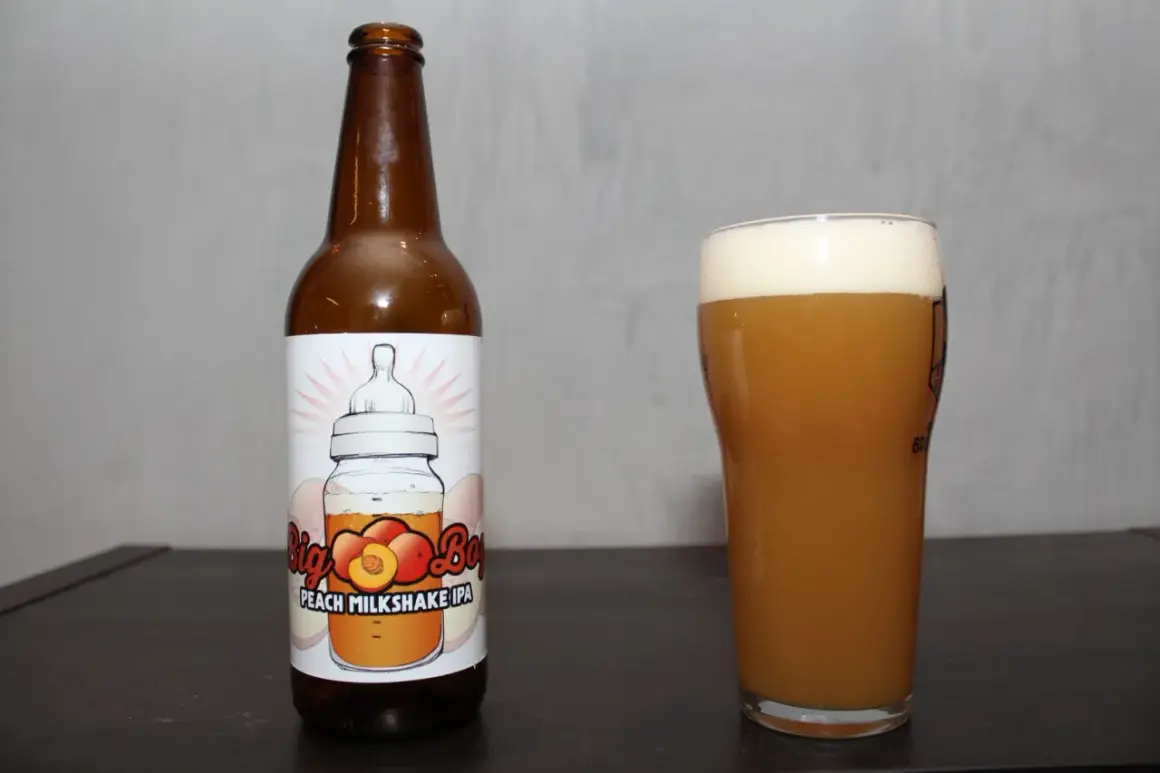 Butelka i szklanka z brzoskwiniowym milkshake IPA. Etykieta przedstawia butelkę z smoczkiem i brzoskwiniami.