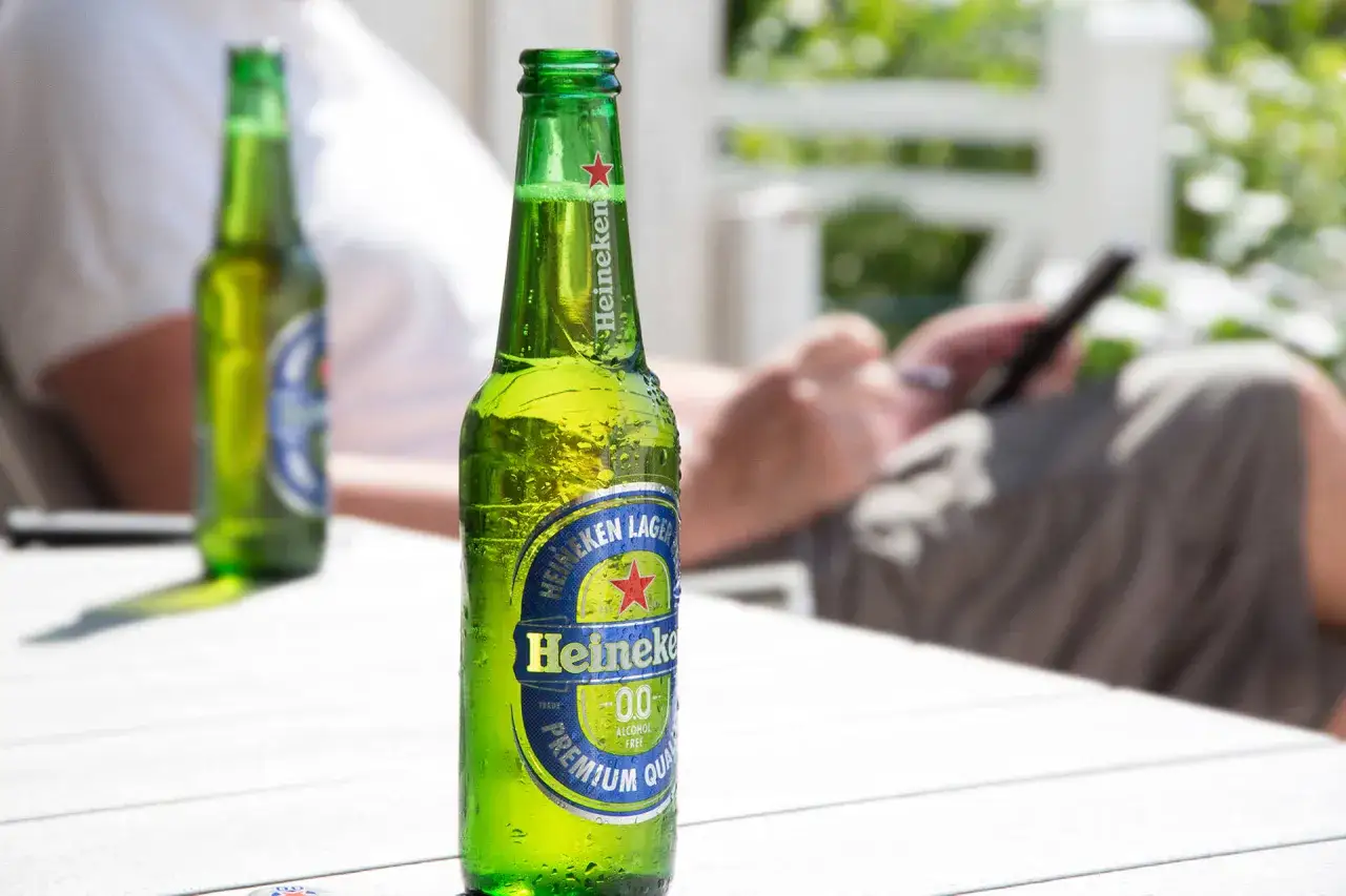 Orzeźwiający alkohol w zielonej butelce Heineken 0.0 na stole, w tle osoba korzystająca z telefonu.