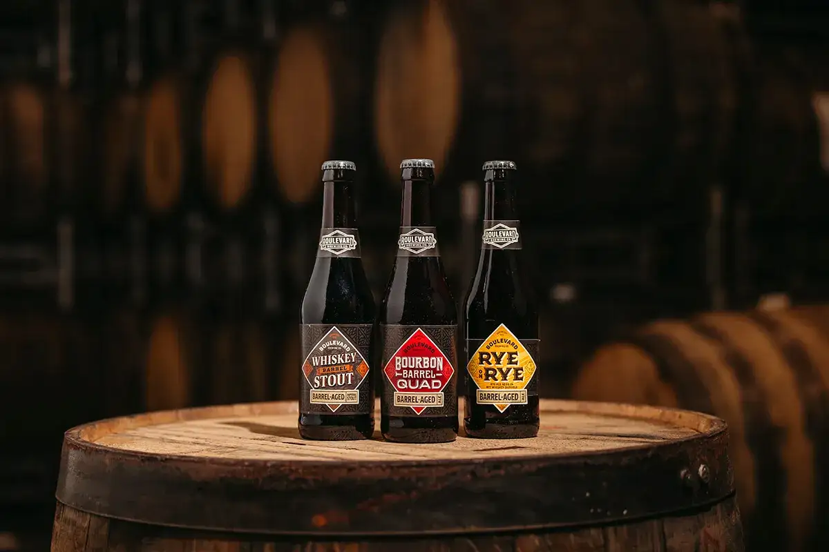 Trzy butelki piwa: Whiskey Stout, Bourbon Barrel Quad i Rye on Rye, wszystkie barrel-aged, stoją na drewnianej beczce.