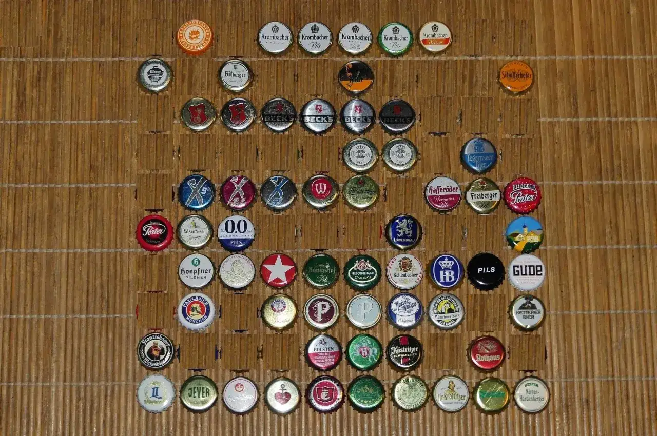 Kolekcja kapsli po piwie z różnych browarów, ułożona na drewnianym tle. Wśród nich widoczne kapsle marek takich jak Bitburger, Beck's, Krombacher, Paulaner, Rothaus i wiele innych.