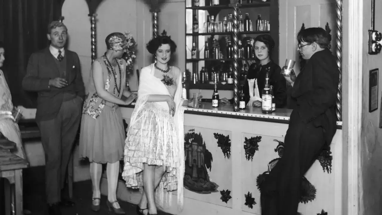 Ludzie przy barze, pijący alkohol. Czy to czasy prohibicji co to? Eleganckie stroje, butelki na półce.