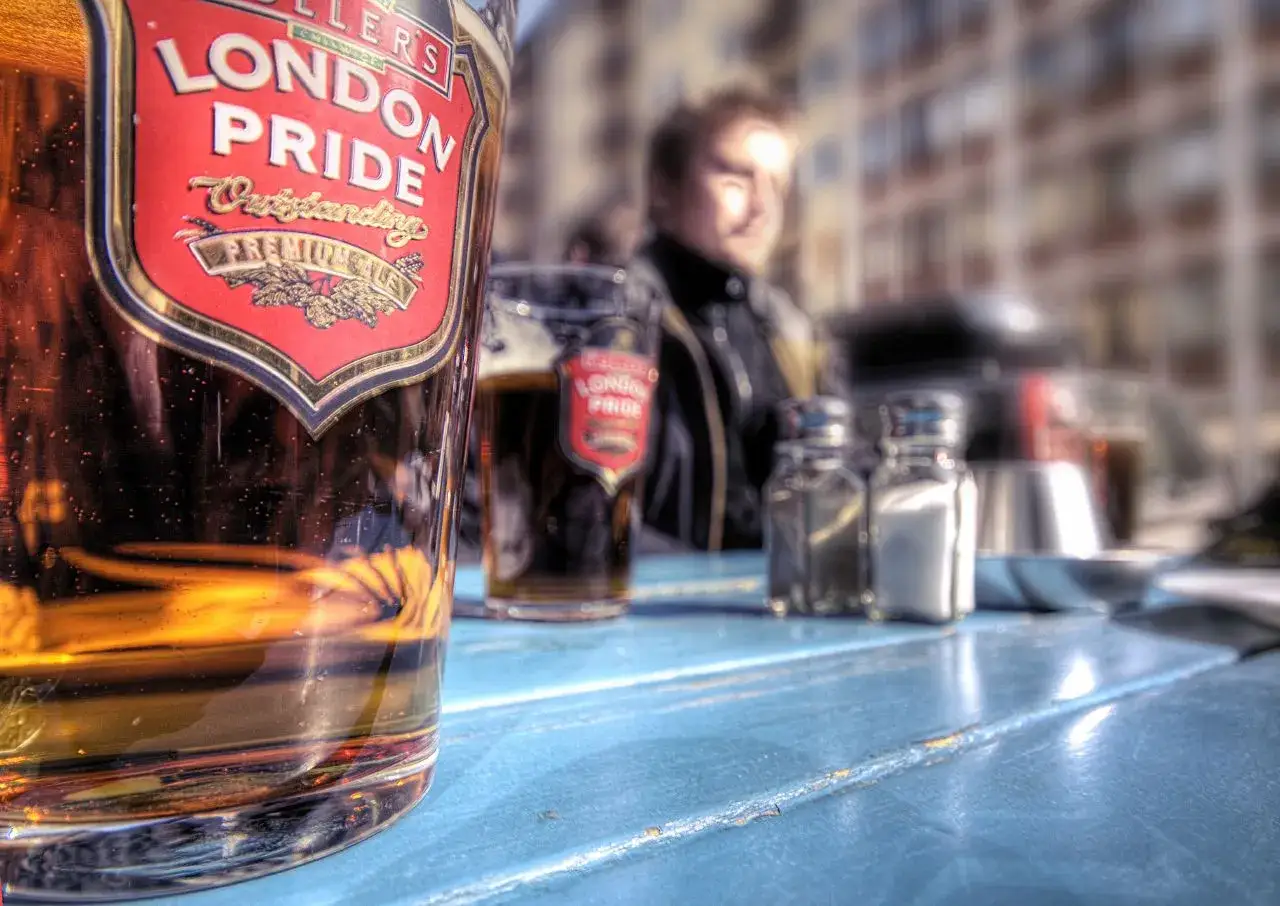 Szklanka z angielskim piwem London Pride na niebieskim stole. W tle widać drugie piwo i sylwetkę mężczyzny.