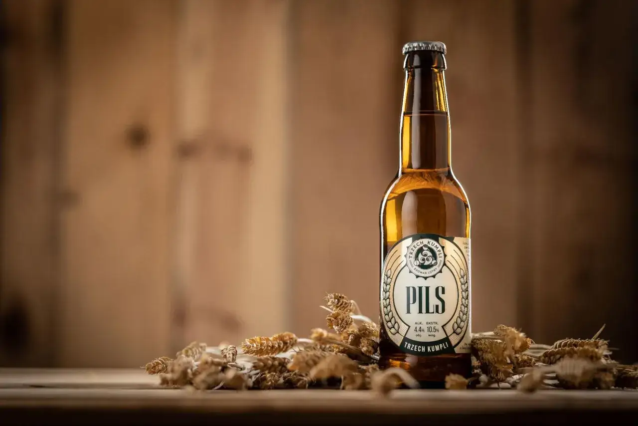 Butelka pils piwo od Trzech Kumpli, otoczona kłosami zb&oacute;ż, na drewnianym tle.