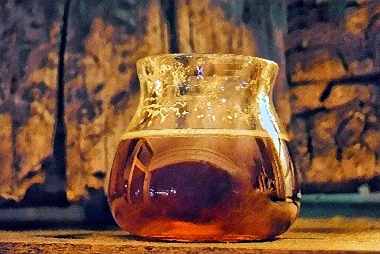 Szklanka z bursztynowym napojem, przypominającym barely wine, stoi na drewnianym blacie.