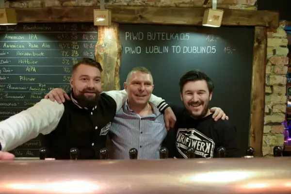 Trzech uśmiechniętych mężczyzn za **tap barem** w pubie. Na tablicy za nimi widnieje menu piwne.