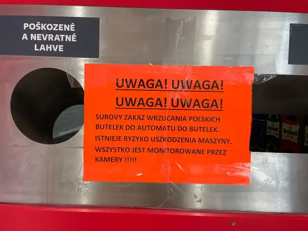 Na automacie do butelek wisi pomarańczowa tabliczka z napisem "UWAGA! UWAGA! SUROWY ZAKAZ WRZUCANIA POLSKICH BUTELEK DO AUTOMATU DO BUTELEK. ISTNIEJE RYZYKO USZKODZENIA MASZYNY. WSZYSTKO JEST MONITOROWANE PRZEZ KAMERY !!!!!". Obok znajduje się czarna t...