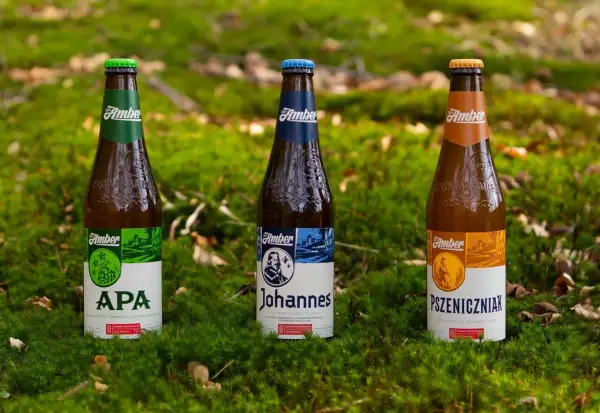 Trzy butelki piwa Amber: APA, Johannes i Pszeniczniak, stoją na mchu. Złote lwy z logo Amber są widoczne na butelkach.