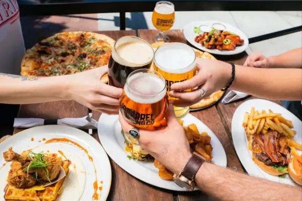 Ludzie wznoszą toasty piwem, celebrując spożycie piwa na świecie przy stole pełnym pizzy, burgerów i kurczaka z goframi.