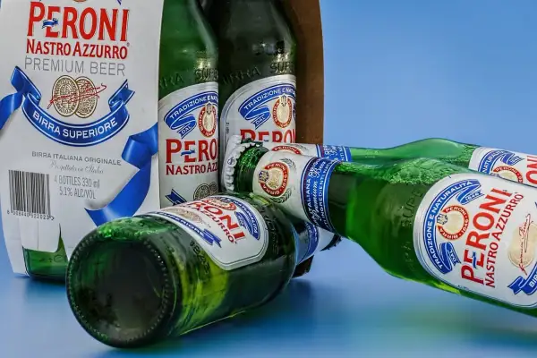 Butelki piwa Peroni Nastro Azzurro leżą na niebieskim tle. Jedna z butelek jest otwarta, a pianka unosi się z szyjki.