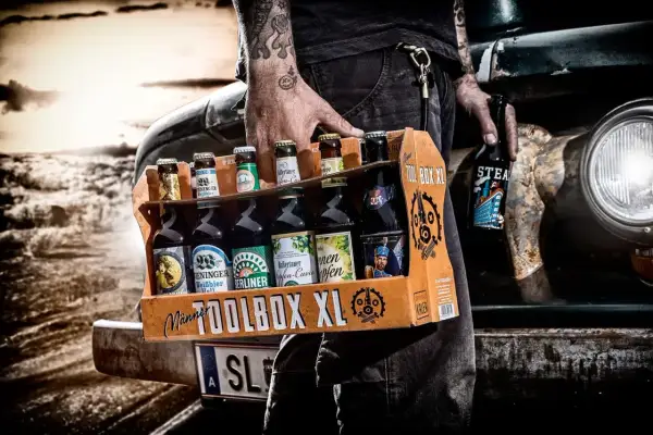 Mężczyzna z tatuażami niesie skrzynkę "TOOLBOX XL" z różnymi butelkami piwa. Idealne połączenie: jakie butelki do jakich skrzynek.